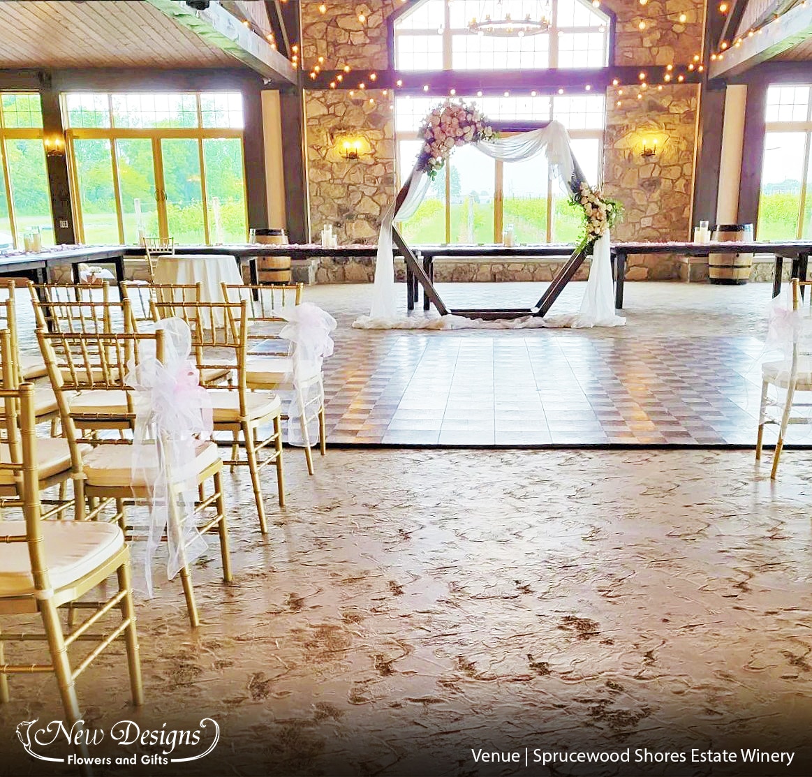 An aisleway in a cobblestone winery, Gold Chiavari Chairs, with a white Organza Bow lining the aisleway, at the end is a Wooden Hexagonal Arch with White draped chiffon, and White and Blush floral installments